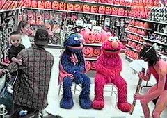 people dressed as Super Grover and Elmo sitting in the Halloween aisle in a store; on the left of the image is a man wearing a baseball cap and holding a baby; on the right of the image is a man in nothing but a loin cloth and a headband