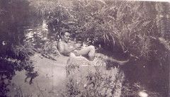 a black and white photo of a man in a body of water next to some plants; the man may or may not be naked