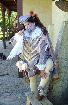 a man in very violet courtly renaissance dress, he is heavily made up and carrying a lace handkerchief