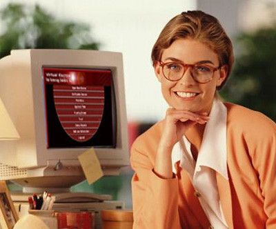 a color photo of an attractive blonde woman with tortoise-shell glasses and a peach-colored blazer; she sits in front of a beige, CRT monitor with a yellow sticky note attached to it