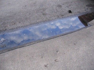 the sky reflected in water in a gutter; a smashed plastic bottle can be seen all the way to the left