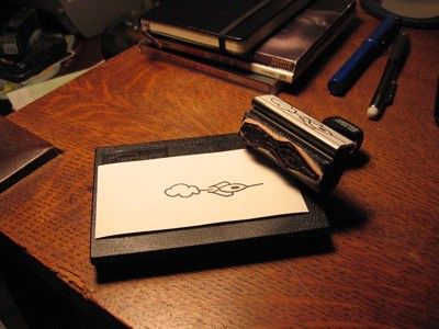 several items on an oak desk: a stamp pad, a stamped piece of paper, a stamp of the rocket to nowhere, a mechanical pencil, a Lamy Safari, 3 CDs, a Moleskine notebook
