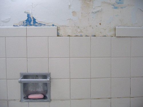 the tiled wall of a bathtub: some paint is peeling, some tiles are missing, the soap in the holder is pink