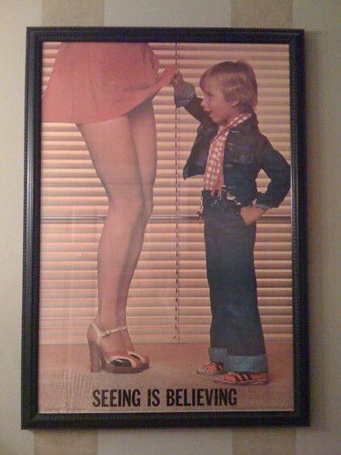 a framed poster of a small boy in a jean jacked, jeans, and orange Adidas shoes lifting the short, red skirt of a long legged woman in very high heels; at the bottom of the poster is the legend "seeing is believing"