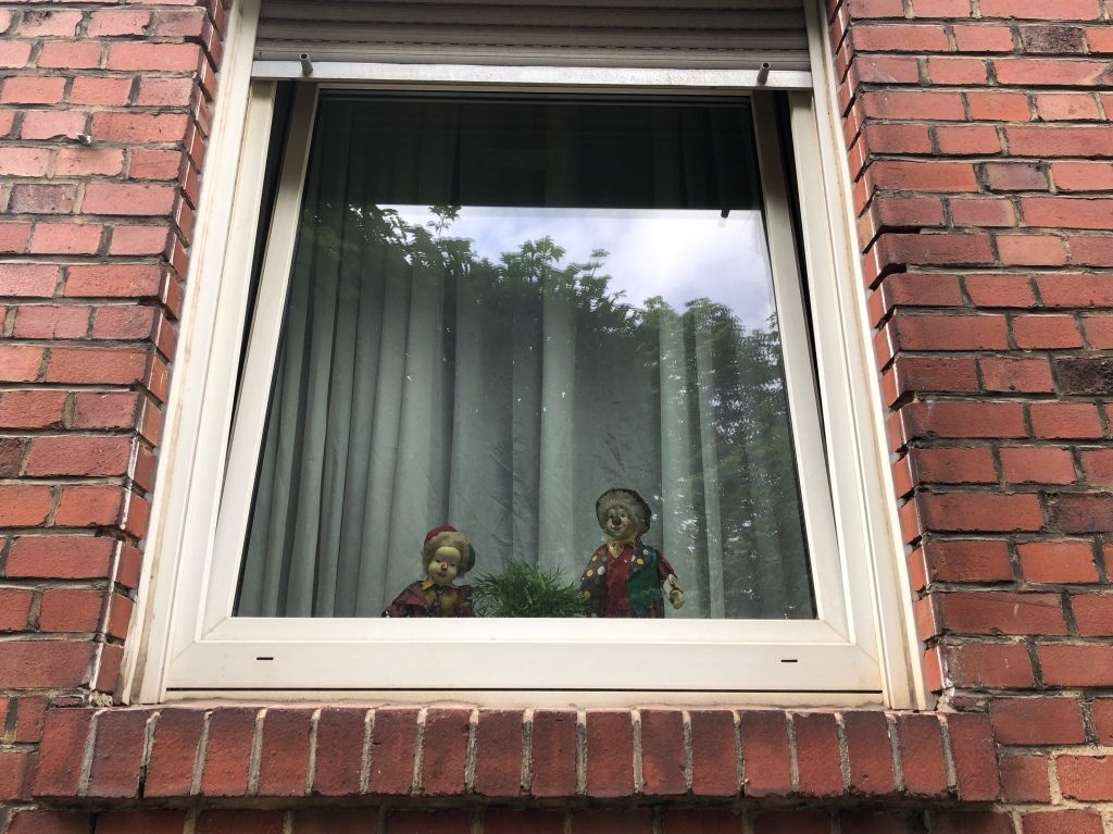 two porcelain clowns behind a window in front of a sheer white curtain; all in a red brick façade