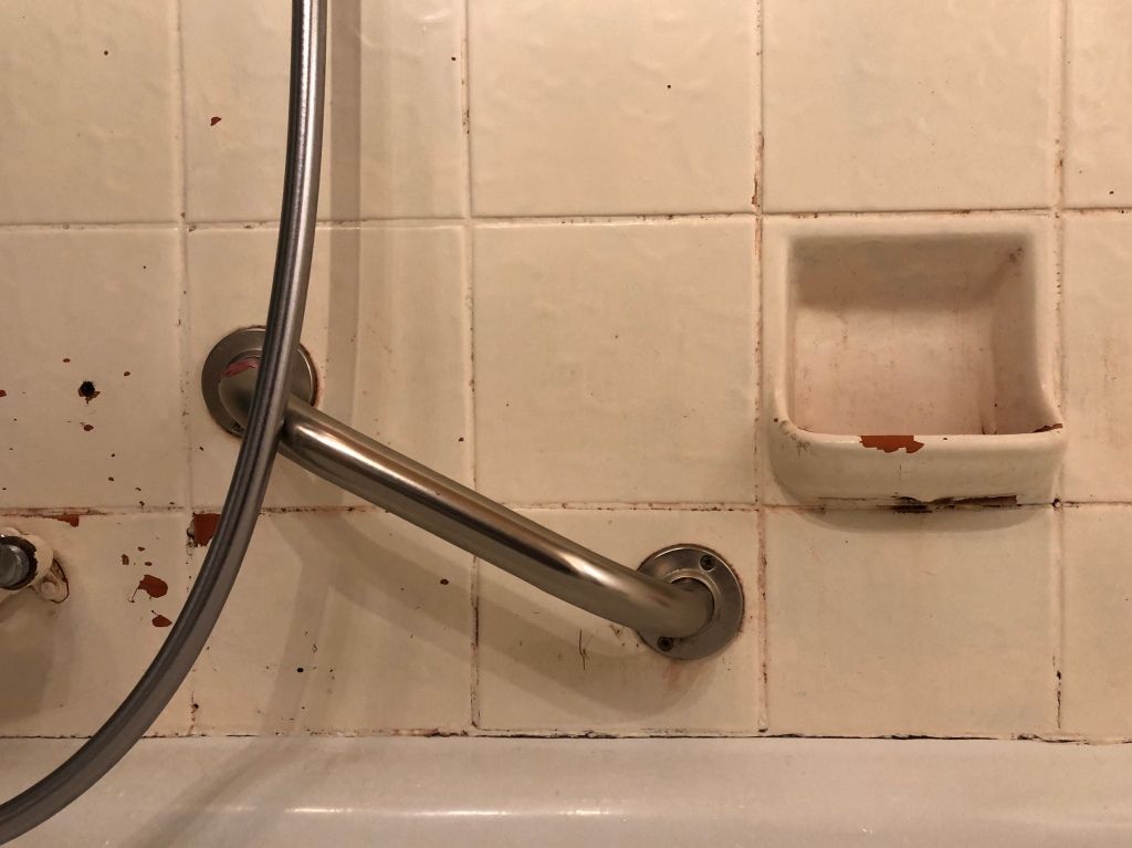 a handle, a silver, plastic hose, and the built-in soap dish in a tiled bathtub where the paint is flaking off