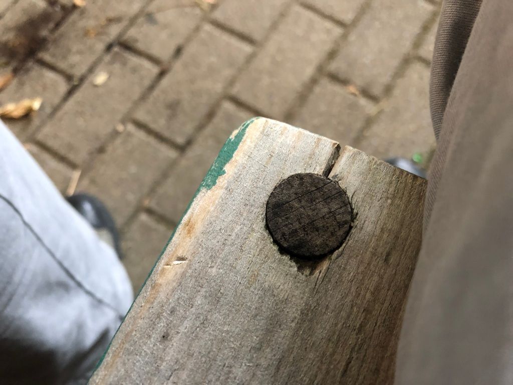 the corner of a wooden bench seat which was once painted green but is now very faded. the visible seat slat is held in place by a large dowel of much darker wood.
