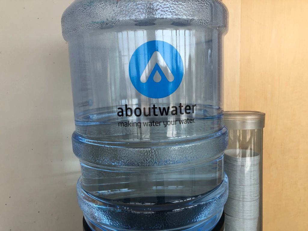 a blue, transparent water jug (in an office setting) upon which has been printed a company's logo, the name of the company, "aboutwater," and the company's slogan, "making water your water."