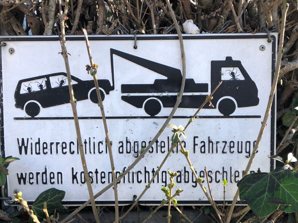 on a chainlink fence with bushes behind it and parts of the bushes in front of it, a black on white metal sign with the legend "Widerrechtliche abgestelte Fahrzeuge werden kostenpflichtig abgeschlept" and above that an icon of a family car attached to a towtruck. black stickers have been placed on the sign to make it look as though both vehicles are being driven by ants