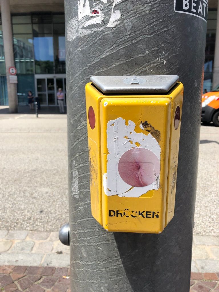 a yellow, German crosswalk pushbutton onto which has been placed a round sticker showing nothing other than a very bleached anus