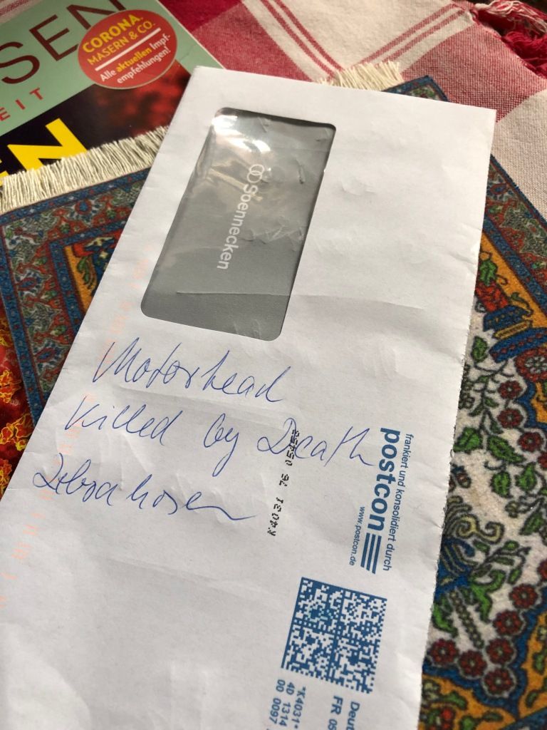 from back to front: a red and white check tablecloth; a partial magazine cover upon which can be seen a red circle containing the words, "Corona, Masern & Co - Alle aktuellen Impf-empfehlungen!; a persian rug mousepad; and a white, plastic window envelope upon which have been written, with a blue, ball-point pen, the words, "Motorhead / Killed by Death / Zebrahosen"