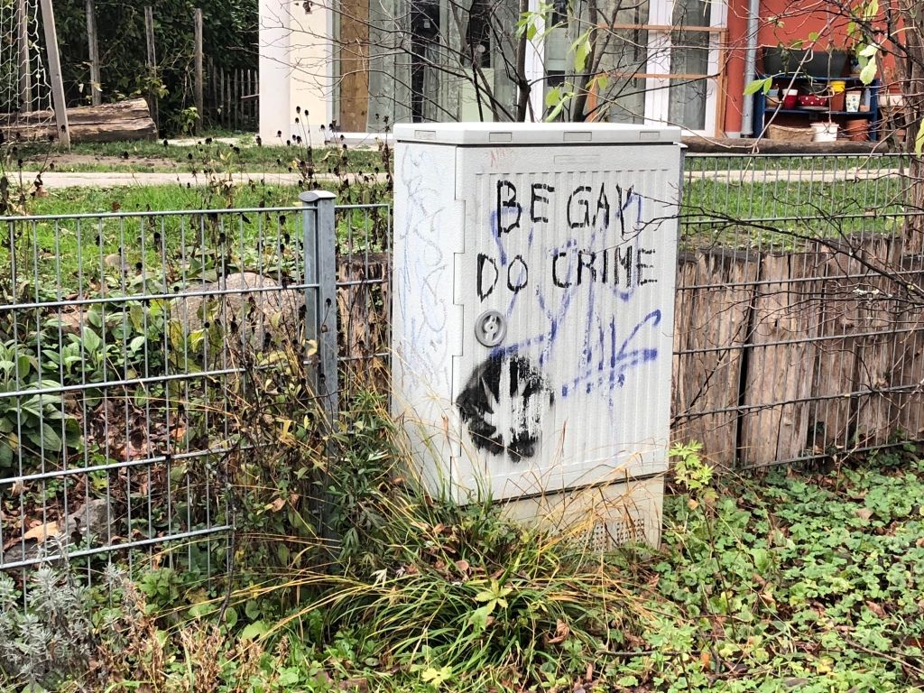 a gray electrical box nestled in a green, autumnal residential scene, and upon which has been graffitied: "BE GAY DO CRIME" and what appears to be a cannabis leaf