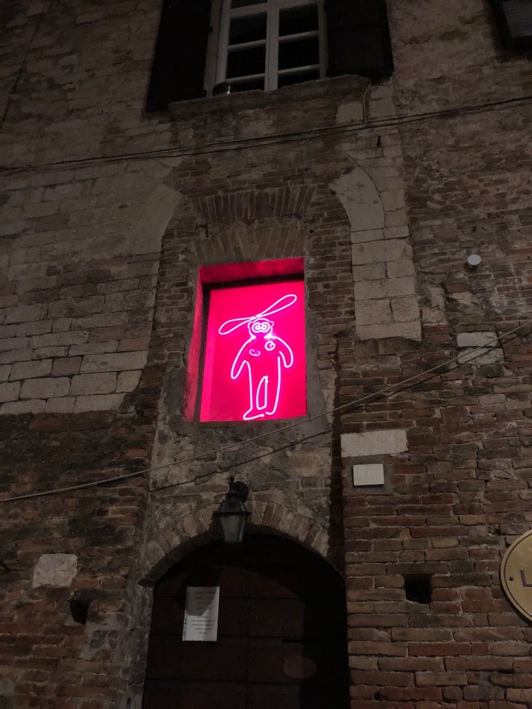 a brick wall which has been changed and repaired several times over the years. above a door in the wall is a window with a bright pink, neon sign in it. the sign is a very simple human figure with nipples, wide eyes, and a very large propeller on its head.