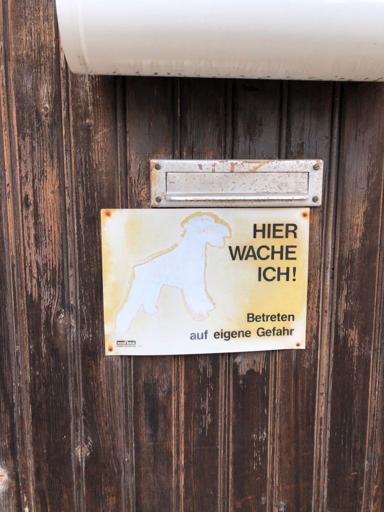 a wooden gate. the stain and varnish on the wood are flaking and fading. at the top, there is a white metal newspaper holder. under the newspaper holder is a metal postal slit. and under the postal slit, there is an old metal sign which reads "HIER WACHE ICH! Betretn auf eigene Gefahr" the sign used to show a picture of a dog, but it has long since faded into white.