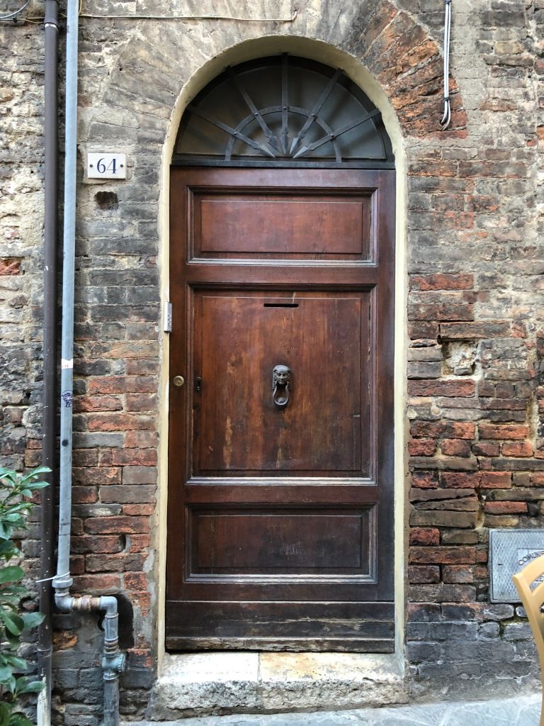 a brick, romanesque arch with a handless, wooden door sporting a look, a post-slit, and an ancient Egypt-inspired knocker; there is a fan-light-shaped metal transom above the door; the house number "64" can also bee seen
