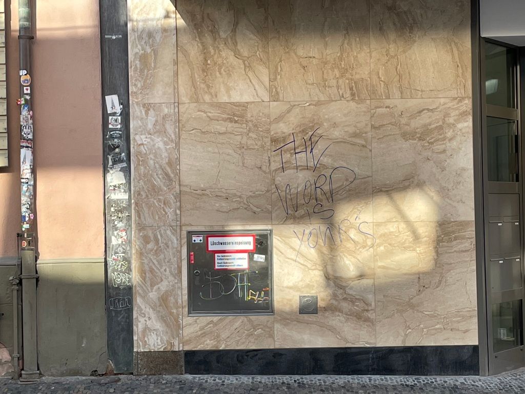 tan marble squares make up the façade of a building into which has been placed a small, metal door upon which a sticker reading "Löschwassereinspeisung" can be found, and onto which has been written with purple permanent marker, "THE WORD IS YOUR'S" 
