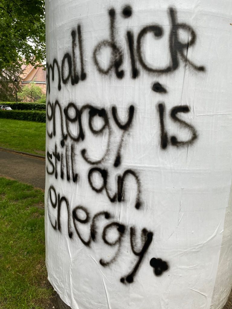 an advertising pillar plastered with white paper upon which has been spray painted in black "small dick energy is still an energy."