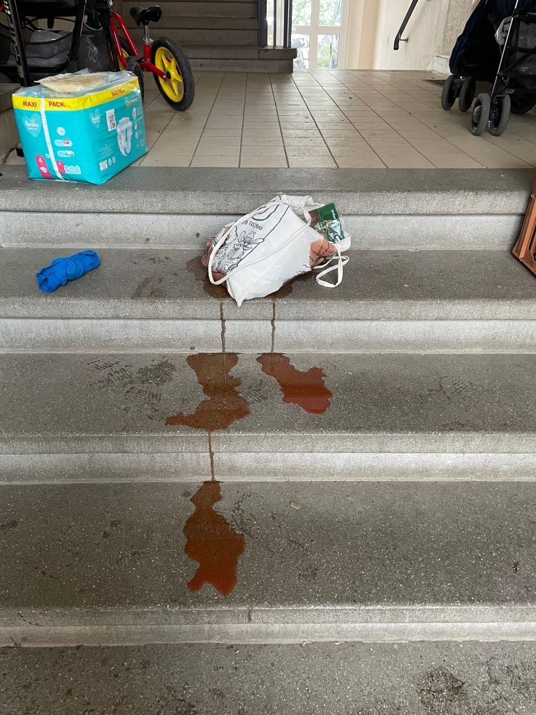 concrete steps in an apartment building's entryway; a blue cotton bag rolled into a package; a pack of diapers; a balance bike; a stroller; a bicycle pump; and, in the middle of it all, a white cotton bag leaking quantities of red liquid