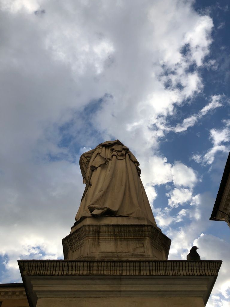 a black pigeon sitting on a stone plinth upon which one can see the back of a statue against a blue and white sky