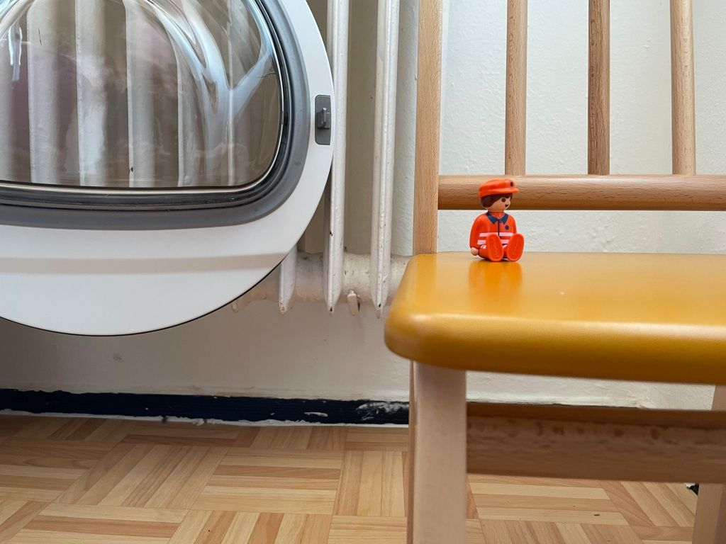 an image of a Playmobil streets and sanitation worker sitting on a mustard yellow child's chair next to an open front-loading washing machine in front of a heating register